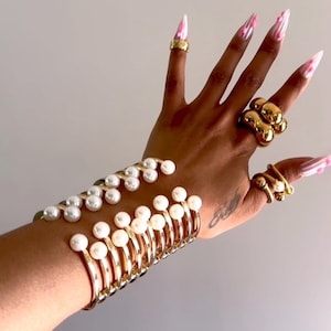 May include: Gold-toned jewelry set featuring a cuff bracelet with white pearls, multiple gold rings, and long, pink and white patterned nails. The jewelry is displayed on a hand against a neutral background.