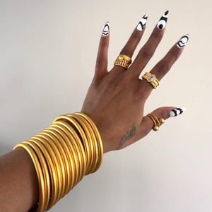 Puede incluir: Una mano adornada con joyas de oro, incluyendo múltiples anillos y una pila de brazaletes. Las uñas son largas y pintadas de blanco con diseños abstractos negros. Las joyas tienen un acabado pulido y reflectante.