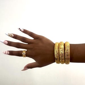 May include: Gold-toned jewelry set against a white background. The image features three ornate bangles, a ring, and a bejeweled nail design. The bangles have detailed patterns. The ring has a square design. The nails are painted with a light pink color.