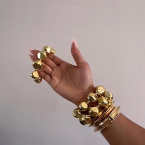 May include: Gold-toned jewelry set featuring a bracelet with large, geometric beads and several stacked bracelets. The bracelets are displayed on a person's arm against a neutral background. The jewelry has a polished finish.