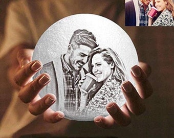 May include: A white moon lamp with a personalized photo of a couple. The photo is engraved on the surface of the lamp, which is illuminated from within. The lamp is being held in a person's hands.