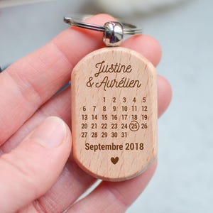 Peut inclure: Un porte-clés en bois avec un calendrier. Il porte les noms "Justine & Aurélien" et un calendrier de septembre 2018, avec le 25 encerclé. Un petit cœur est en bas.