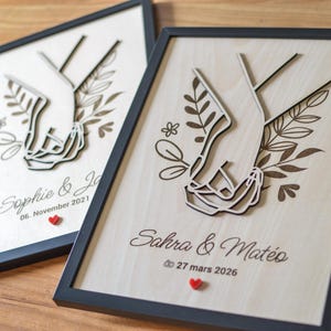 Puede incluir: Dos obras de arte enmarcadas en madera con un dise&ntilde;o cortado con l&aacute;ser de manos entrelazadas. Cada pieza incluye nombres, una fecha y un peque&ntilde;o coraz&oacute;n rojo. Los marcos son negros.