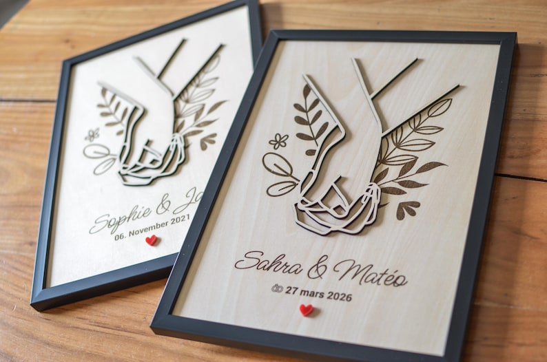 Puede incluir: Dos obras de arte enmarcadas en madera con un dise&ntilde;o cortado con l&aacute;ser de manos entrelazadas. Cada pieza incluye nombres, una fecha y un peque&ntilde;o coraz&oacute;n rojo. Los marcos son negros.