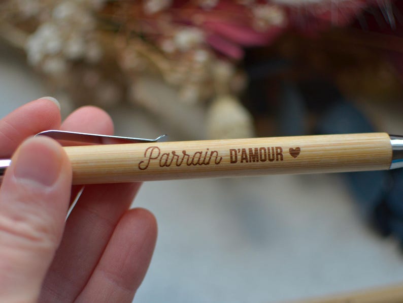 Op de afbeelding: Een houten pen met een zilveren clip en de woorden "Parrain D'Amour" erop gegraveerd. Een klein hartje is ook op de pen gegraveerd.