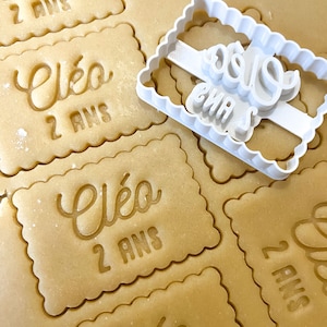 Peut inclure: Gros plan sur un emporte-pièce blanc avec le mot "Eléo" et "2 ans" en cursive. L'emporte-pièce est posé sur une feuille de pâte à biscuits avec plusieurs autres biscuits déjà découpés avec le même motif.