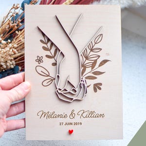Puede incluir: Placa de madera con dise&ntilde;o cortado con l&aacute;ser de dos manos entrelazadas, rodeadas de elementos florales. Los nombres "M&eacute;lanie & Killian" y la fecha "27 JUIN 2019" est&aacute;n grabados debajo, con un peque&ntilde;o coraz&oacute;n rojo. Un regalo &uacute;nico de boda o aniversario.