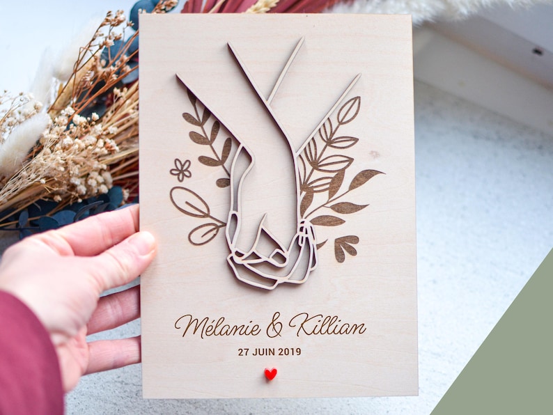 Puede incluir: Placa de madera con dise&ntilde;o cortado con l&aacute;ser de dos manos entrelazadas, rodeadas de elementos florales. Los nombres "M&eacute;lanie & Killian" y la fecha "27 JUIN 2019" est&aacute;n grabados debajo, con un peque&ntilde;o coraz&oacute;n rojo. Un regalo &uacute;nico de boda o aniversario.