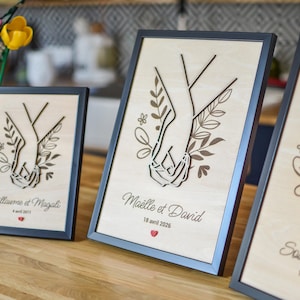 Puede incluir: Tres obras de arte enmarcadas en madera con un dise&ntilde;o cortado con l&aacute;ser de dos manos entrelazadas, rodeadas de elementos florales. Cada marco tiene un borde negro y texto personalizado con fechas. Las obras se exhiben sobre una superficie de madera.