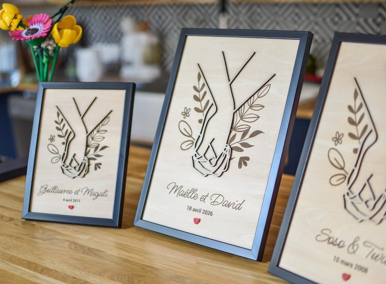 Puede incluir: Tres obras de arte enmarcadas en madera con un dise&ntilde;o cortado con l&aacute;ser de dos manos entrelazadas, rodeadas de elementos florales. Cada marco tiene un borde negro y texto personalizado con fechas. Las obras se exhiben sobre una superficie de madera.