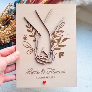 Puede incluir: Un recuerdo de boda de madera con manos entrelazadas cortadas con l&aacute;ser, rodeadas de elementos florales. Los nombres "Lucie & Flavien" y la fecha "1 OCTOBRE 2012" est&aacute;n grabados, con un peque&ntilde;o coraz&oacute;n rojo. Fondo de madera clara.