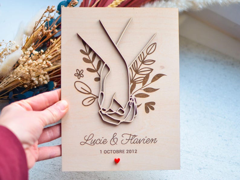 Puede incluir: Un recuerdo de boda de madera con manos entrelazadas cortadas con l&aacute;ser, rodeadas de elementos florales. Los nombres "Lucie & Flavien" y la fecha "1 OCTOBRE 2012" est&aacute;n grabados, con un peque&ntilde;o coraz&oacute;n rojo. Fondo de madera clara.