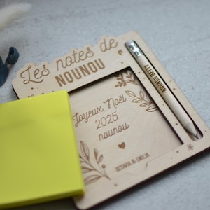 Peut inclure: Tableau mémo en bois avec un crayon et un bloc-notes jaune. Le tableau porte des inscriptions gravées en français, dont "Les notes de NOUNOU" et "Joyeux Noël 2025 nounou". Un crayon avec du texte est dans le support.