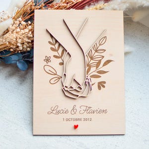Puede incluir: Placa de boda de madera con un dise&ntilde;o cortado con l&aacute;ser de dos manos entrelazadas, rodeadas de detalles florales. Los nombres "Lucie & Flavien" y la fecha "1 Octobre 2012" est&aacute;n grabados, con un peque&ntilde;o coraz&oacute;n rojo.
