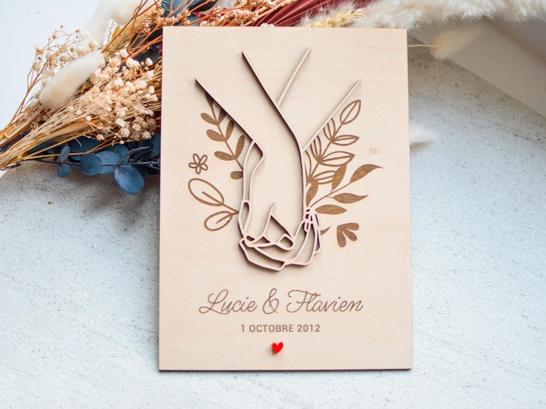 Puede incluir: Placa de boda de madera con un dise&ntilde;o cortado con l&aacute;ser de dos manos entrelazadas, rodeadas de detalles florales. Los nombres "Lucie & Flavien" y la fecha "1 Octobre 2012" est&aacute;n grabados, con un peque&ntilde;o coraz&oacute;n rojo.