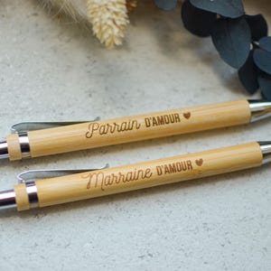 Op de afbeelding: Twee houten pennen met zilveren accenten. De pennen zijn gegraveerd met de woorden "Parrain d'Amour" en "Marraine d'Amour" met een klein hartje na elke zin.