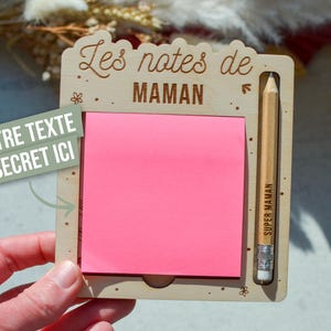 Peut inclure: Tableau mémo en bois avec un bloc-notes rose et un crayon. Le tableau porte l'inscription "Les notes de MAMAN" et "SUPER MAMAN" sur le crayon. Une boîte de texte verte indique "VOTRE TEXTE SECRET ICI".