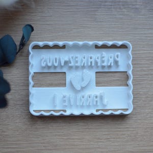 Peut inclure: Un emporte-pièce en plastique blanc avec un bord festonné. Le texte "PRÉPAREZ-VOUS ! ARRIVE 1" est en relief sur l'emporte-pièce, ainsi qu'une paire de pieds de bébé. L'emporte-pièce est sur une surface en bois.