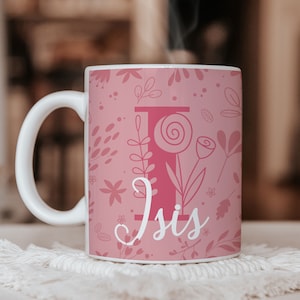 Peut inclure: Une tasse en céramique rose avec un motif floral et la lettre "I" en rose. Le nom "Isis" est écrit en cursive sous la lettre.