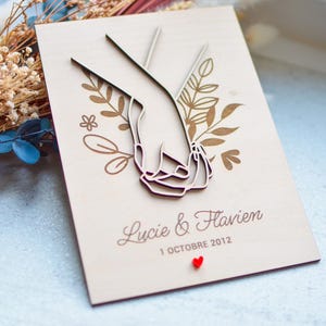 Puede incluir: Un recuerdo de boda de madera con un dise&ntilde;o cortado con l&aacute;ser de dos manos entrelazadas, rodeado de elementos florales. Los nombres "Lucie & Flavien" y la fecha "1 Octubre 2012" est&aacute;n grabados debajo, con un peque&ntilde;o coraz&oacute;n rojo.