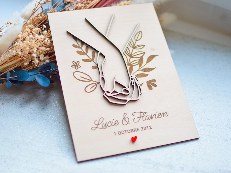 Puede incluir: Un recuerdo de boda de madera con un dise&ntilde;o cortado con l&aacute;ser de dos manos entrelazadas, rodeado de elementos florales. Los nombres "Lucie & Flavien" y la fecha "1 Octubre 2012" est&aacute;n grabados debajo, con un peque&ntilde;o coraz&oacute;n rojo.