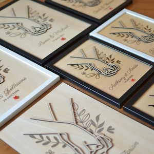 Puede incluir: Obras de arte de madera cortadas con l&aacute;ser que presentan manos entrelazadas, detalles florales y texto personalizado, enmarcadas en blanco y negro. Ideal para regalos de boda o aniversario.