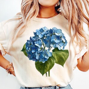 Könnte beinhalten: Cremefarbenes T-Shirt mit einem blauen Hortensien-Blumenmuster. Die Grafik zeigt detaillierte blaue Blumencluster, grüne Blätter und Stiele. Das Shirt besteht aus einem weichen Material und hat hochgekrempelte Ärmel.