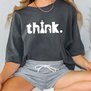 Könnte beinhalten: Dunkelgraues T-Shirt mit dem Wort "think" in Weiß, mit Schachfiguren-Silhouetten. Das Shirt wird mit grauen Shorts kombiniert. Das Wort "Pepper" befindet sich in der oberen rechten Ecke.