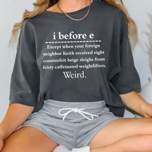 Könnte beinhalten: Dunkelgraues T-Shirt mit dem Text "i before e" und einer humorvollen Ausnahme. Das Shirt enthält auch das Wort "Weird". Das Model trägt graue Shorts und weiße Socken.
