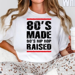 Könnte beinhalten: Ein weißes kurzärmeliges T-Shirt mit schwarzem, abgenutztem Text, der 80'S MADE 90'S HIP HOP RAISED lautet, eingerahmt von zwei horizontalen roten Linien. Das T-Shirt wird mit einer hellen Hose mit braun-schwarzem Leopardenmuster kombiniert.