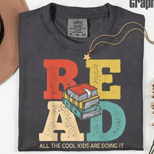 Könnte beinhalten: Graphitfarbenes T-Shirt mit dem Wort "READ" in bunten, abgenutzten Buchstaben, einer Grafik von gestapelten Büchern und dem Satz "ALL THE COOL KIDS ARE DOING IT". Eine goldene Halskette mit Sternanhänger liegt auf dem T-Shirt.