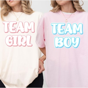 May include: Two short-sleeved t-shirts, one cream and one pink. The cream shirt has the text "TEAM GIRL" in pink bubble letters. The pink shirt has the text "TEAM BOY" in blue bubble letters. Both shirts are worn by a person.