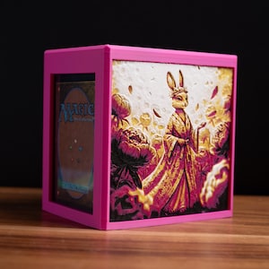 Op de afbeelding: Een felroze kaartenopbergdoos met een gedetailleerde illustratie van een konijn in een gewaad. De doos bevat een kaart met de tekst "Magic The Gathering". De doos staat op een houten oppervlak.