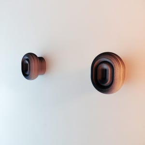 Puede incluir: Dos pomos de madera para armario con una combinación de colores marrón oscuro y beige. Los pomos tienen forma redondeada con un centro ovalado empotrado. Uno está montado en una pared blanca, el otro está a la izquierda.
