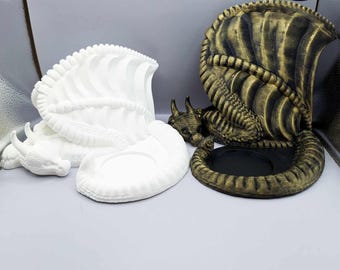May include: Two dragon candle holders, one white and one gold. Both are sculpted in a curled up position with wings spread out. The candle holders have a circular indentation for a candle.