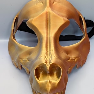 Puede incluir: Una máscara dorada impresa en 3D con forma de cráneo de animal estilizado. La máscara tiene aberturas para los ojos y la nariz, y una correa negra. El cráneo presenta dientes prominentes y una superficie texturizada.