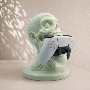 以下が含まれることがあります： 淡い緑色の頭蓋骨型ゲームコントローラーホルダー。このホルダーは、白と黒のゲームコントローラーを保持するように設計されています。頭蓋骨には詳細な特徴があり、丸いベースに座っています。コントローラーは頭蓋骨の口の中に保持されています。