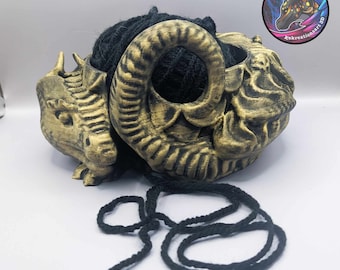 May include: A gold-colored dragon yarn bowl with a black yarn ball inside. The bowl is shaped like a dragon's head with a large horn on the side. The yarn is hanging out of the bowl and is looped around the horn.