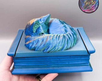 May include: A blue 3D printed box with a sleeping cat on the lid. The cat is curled up and has a green and blue color scheme. The box has a textured surface and a decorative design.