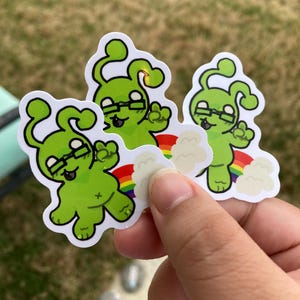 May include: Three green alien stickers with black glasses and a rainbow. The aliens have a cartoonish design with a cross on their belly. The stickers are held by a hand against a blurred green background.