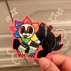 Op de afbeelding: Een kleurrijke sticker met een cartoonfiguur met een bloemvormige kop en regenboogaccenten. Het figuur heeft een zwart lichaam, rode ogen en een groene tong. De sticker wordt door een hand vastgehouden.
