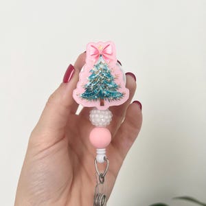 May include: A pink Christmas tree badge reel with a teal tree design, a pink bow, and white and pink beads. The badge reel has a clear plastic clip and a silver-colored metal clasp.