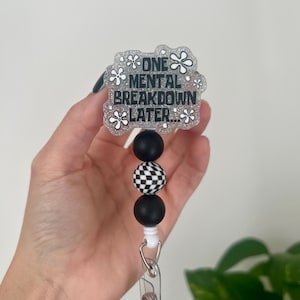 May include: A retractable badge reel with the text "ONE MENTAL BREAKDOWN LATER..." in black on a glittery white background. The reel features black and checkered beads.