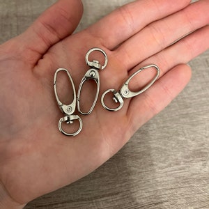 May include: Three silver-colored swivel clasps with oval-shaped openings and circular rings. The clasps are shown on a light-colored surface. These clasps are often used for keychains, lanyards, or bag straps.