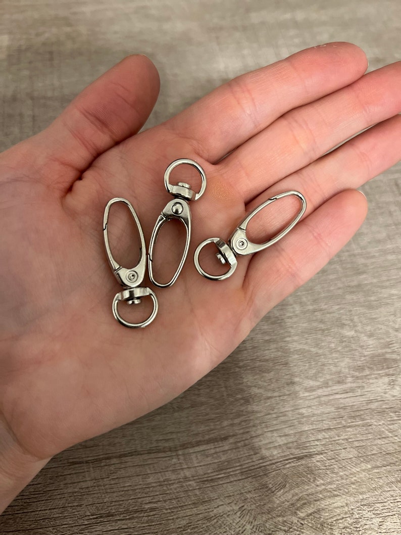 May include: Three silver-colored swivel clasps with oval-shaped openings and circular rings. The clasps are shown on a light-colored surface. These clasps are often used for keychains, lanyards, or bag straps.