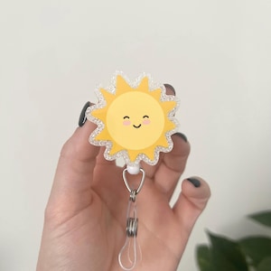 May include: A yellow, smiling sun-shaped badge reel with a glittery, silver border. The badge reel has a retractable cord and a metal clip. The sun has a happy face and is a cheerful accessory.