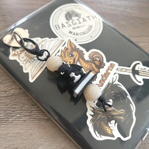 May include: A black and white dragon keychain with two speckled beige beads. The keychain is attached to a clear phone case decorated with stickers, including one that says "BASGIATH WAR COLLEGE".