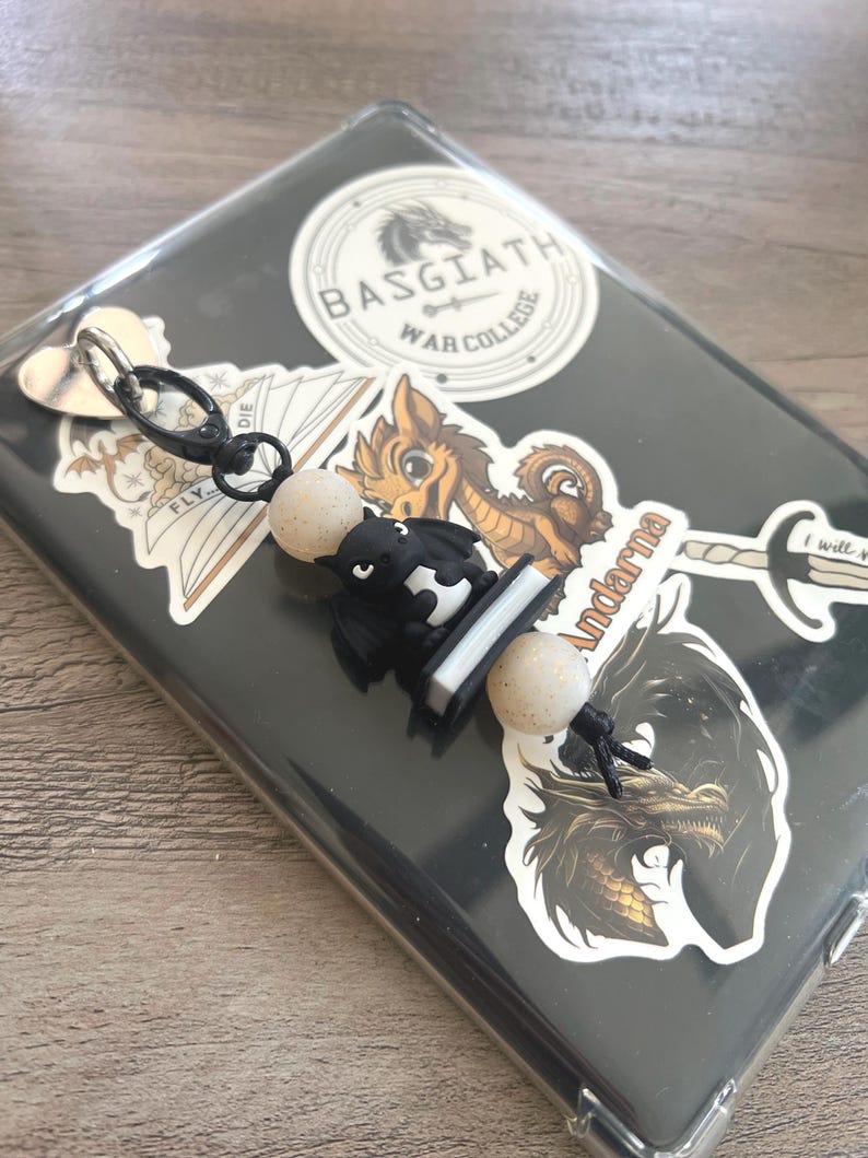 May include: A black and white dragon keychain with two speckled beige beads. The keychain is attached to a clear phone case decorated with stickers, including one that says "BASGIATH WAR COLLEGE".