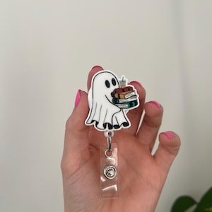 May include: White ghost cartoon character holding a stack of books and a cup of coffee. The ghost is attached to a clear plastic retractable badge holder with a silver button.
