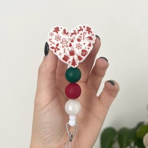 May include: A Christmas-themed badge reel with a heart-shaped design. The white heart features red holiday icons. Below the heart are green, red, and white beads. The reel has a clear plastic strap and a metal clip.